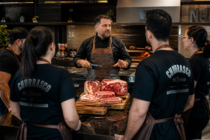 Instrutor ensinando técnicas de churrasco para alunos em cozinha gourmet, cortes de carne expostos, churrasqueira ao fundo, aventais profissionais, estilo fotográfico realista, ambiente premium